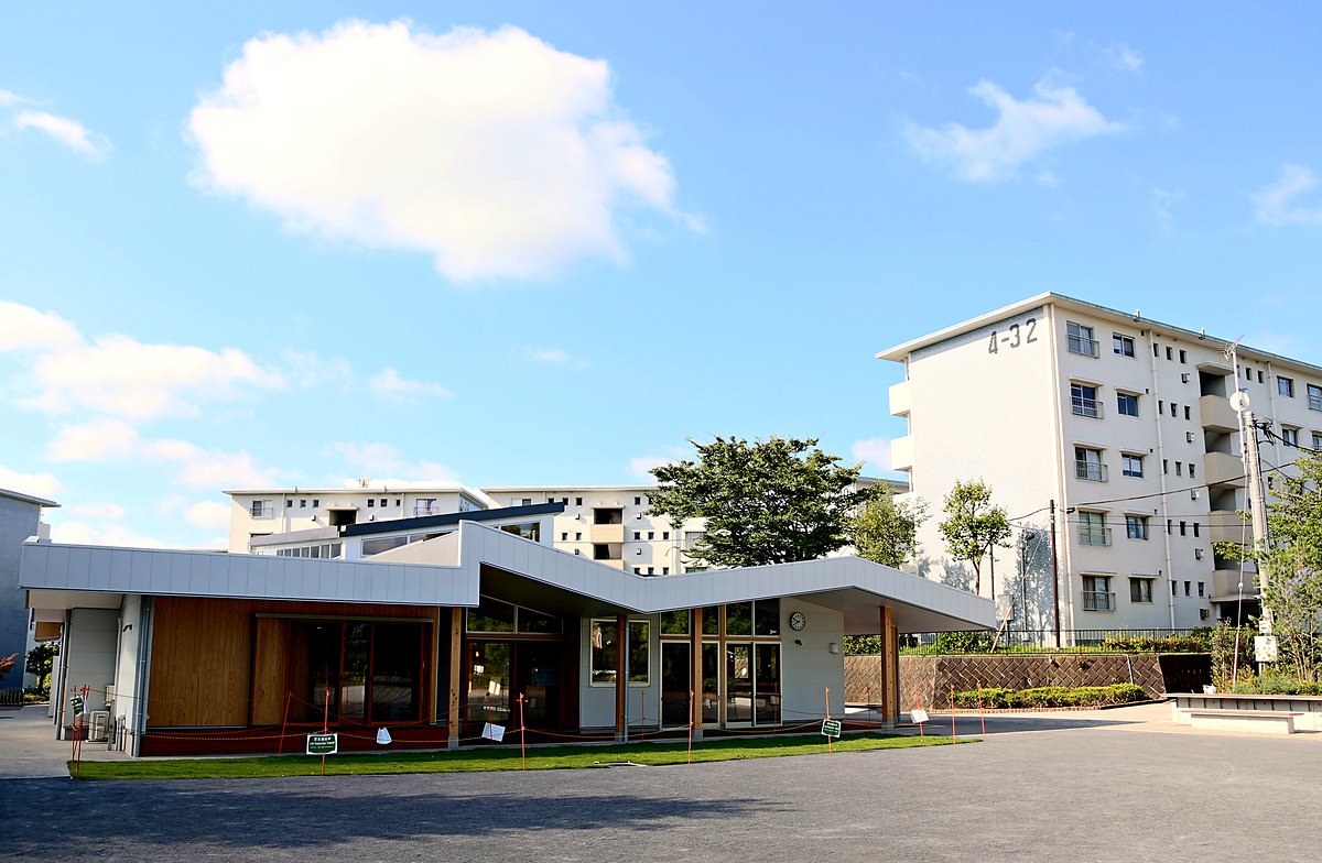 正面から見た木造の集会所。折れ屋根が連なり、背後に団地の住棟